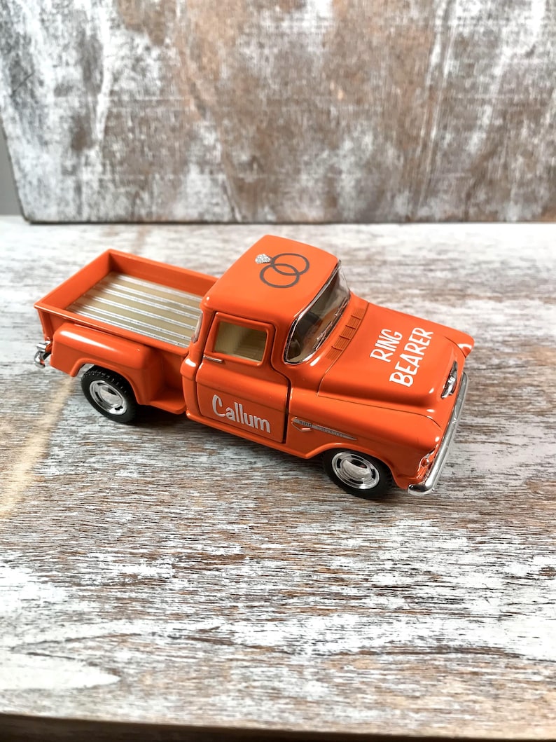 May include: An orange toy pickup truck with the words "Ring Bearer" and "Callum" printed on it. The truck has a silver bed and wheels, and a graphic of two wedding rings on the roof.