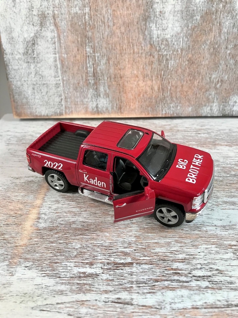 May include: Red toy pickup truck with a black bed and a black interior. The truck has a white sticker on the side that says "BIG BROTHER" and another white sticker that says "Kaden". The truck also has a white sticker on the back that says "2022".