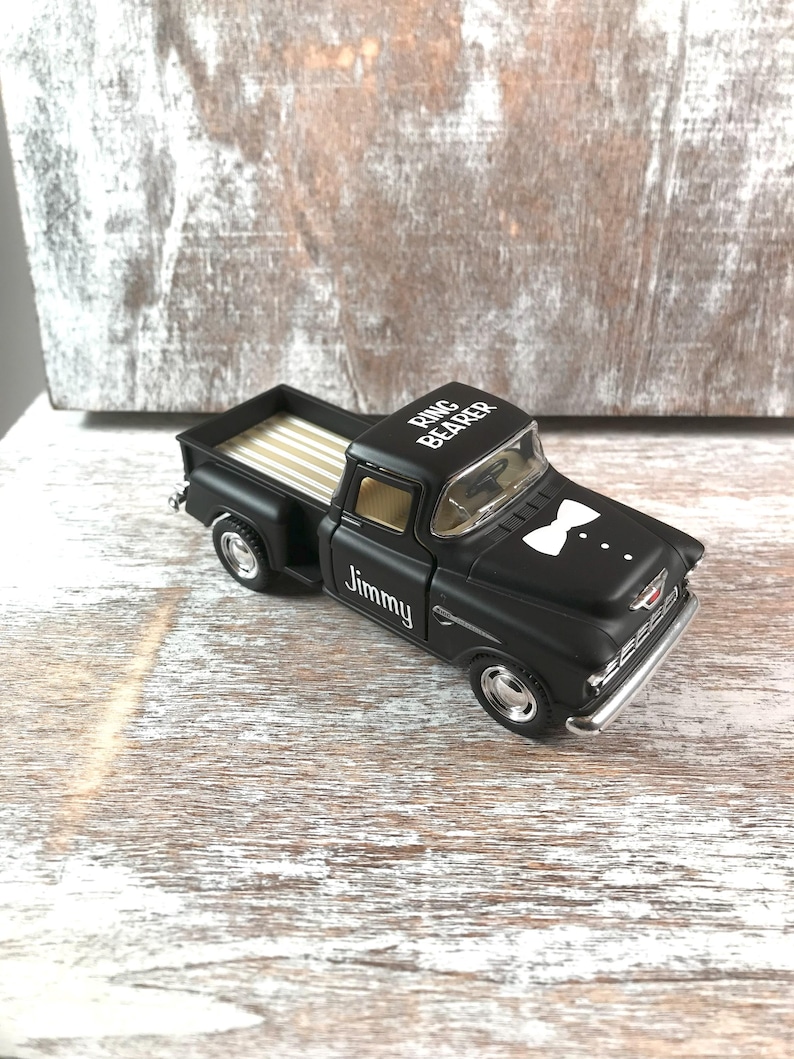 May include: A black toy pickup truck with the words "Ring Bearer" on the roof and "Jimmy" on the side. The truck has a white bow tie graphic on the front and a tan bed.