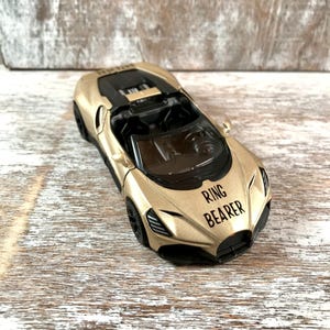 May include: A gold toy car with black accents and the text "RING BEARER" on the front. The car has a sleek design with visible interior details. The toy car is a miniature replica of a sports car.