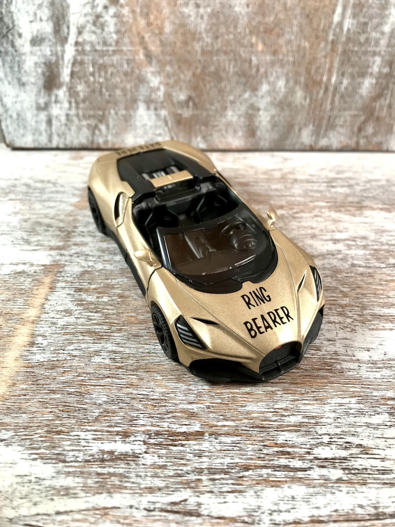 May include: A gold toy car with black accents and the text "RING BEARER" on the front. The car has a sleek design with visible interior details. The toy car is a miniature replica of a sports car.