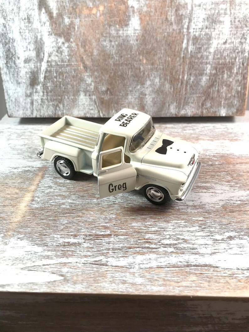 May include: A white toy truck with the words "Ring Bearer" on the roof and "Greg" on the door. The truck has a black bow tie graphic and three dots. The truck is on a wooden surface.