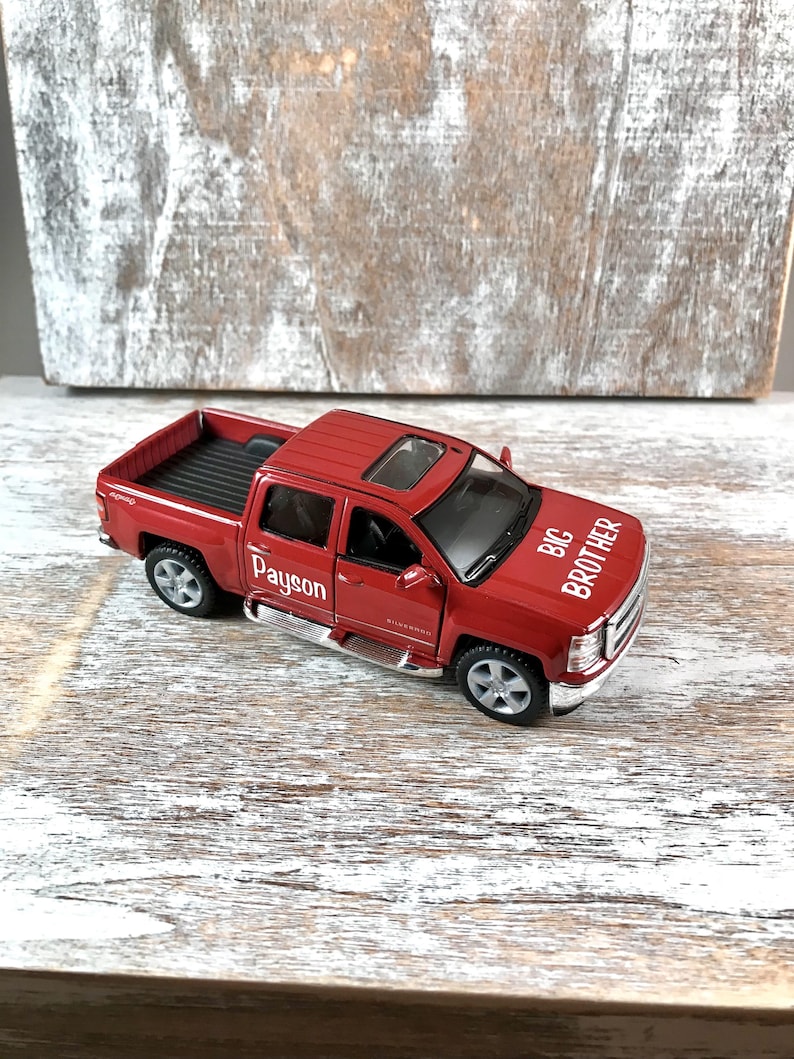 May include: A red toy pickup truck with the words "BIG BROTHER" and "Payson" printed on it. The truck has a sunroof, chrome accents, and silver wheels. The truck is sitting on a light-colored wooden surface.