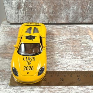 Puede incluir: Un coche de juguete amarillo con el texto "CLASS OF 2026" y "DATE HERE". El coche tiene un gorro de graduaci&oacute;n en la parte superior. Una regla est&aacute; al lado del coche. El coche mide aproximadamente 10 cm de largo.