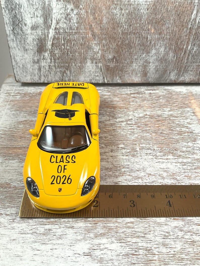 Puede incluir: Un coche de juguete amarillo con el texto "CLASS OF 2026" y "DATE HERE". El coche tiene un gorro de graduaci&oacute;n en la parte superior. Una regla est&aacute; al lado del coche. El coche mide aproximadamente 10 cm de largo.