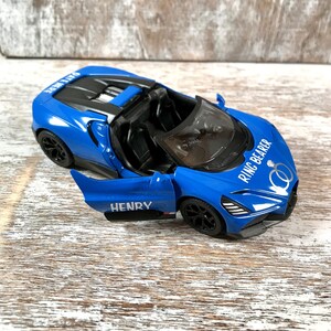 May include: A blue toy convertible car with the door open, featuring the name "HENRY" on the door. The car has black wheels and the words "RING BEARER" and wedding rings are printed on the hood.