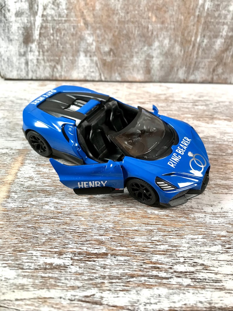 May include: A blue toy convertible car with the door open, featuring the name "HENRY" on the door. The car has black wheels and the words "RING BEARER" and wedding rings are printed on the hood.