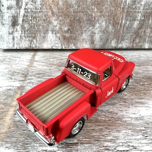 May include: A red toy pickup truck with the text "SECURITY" on the hood and "Jack" on the side. The truck bed has a striped design. The date "3-11-23" is visible on the windshield.
