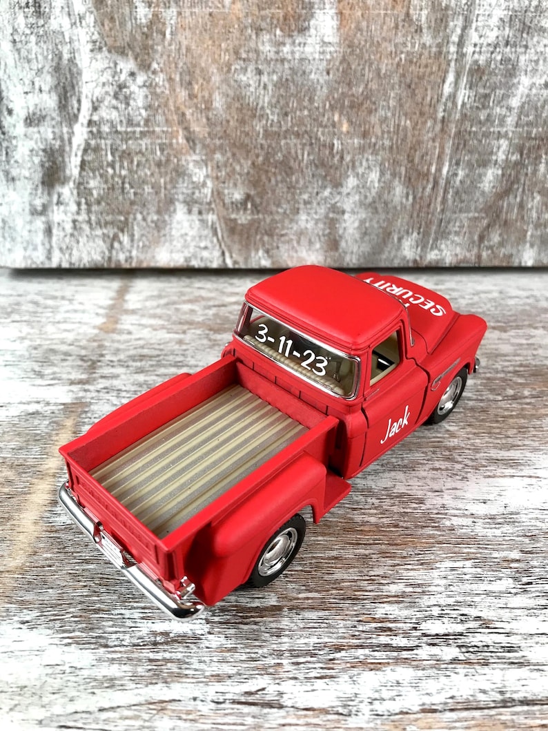 May include: A red toy pickup truck with the text "SECURITY" on the hood and "Jack" on the side. The truck bed has a striped design. The date "3-11-23" is visible on the windshield.