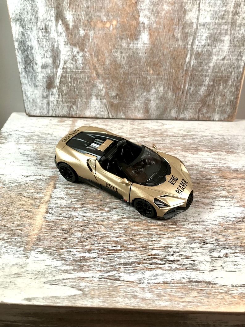 May include: A gold-colored toy convertible car with black accents. The car has the words "RING BEARER" and "KYLE" printed on it. The car is sitting on a white and gray wooden surface.