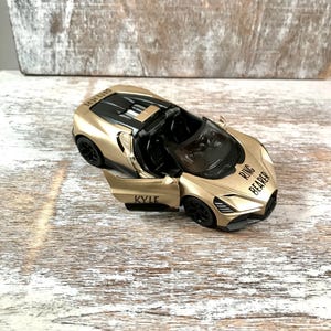 May include: A gold-colored toy sports car with the door open, revealing a black interior. The car has black wheels and the text "RING BEARER" and "KYLE" printed on it. The car is on a light-colored wooden surface.