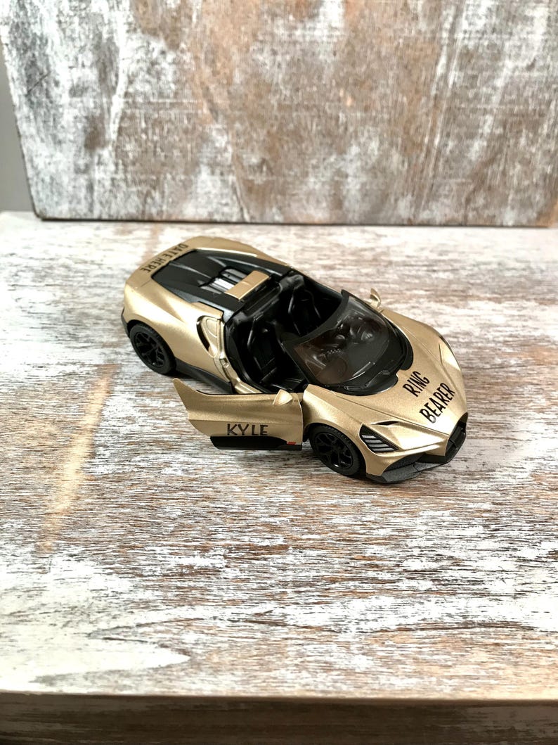 May include: A gold-colored toy sports car with the door open, revealing a black interior. The car has black wheels and the text "RING BEARER" and "KYLE" printed on it. The car is on a light-colored wooden surface.