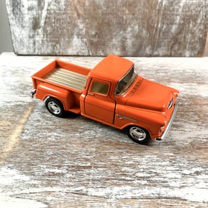 May include: An orange vintage pickup truck model with silver accents. The truck has a detailed design, including a bed with silver stripes and chrome wheels. The model is a classic collectible.