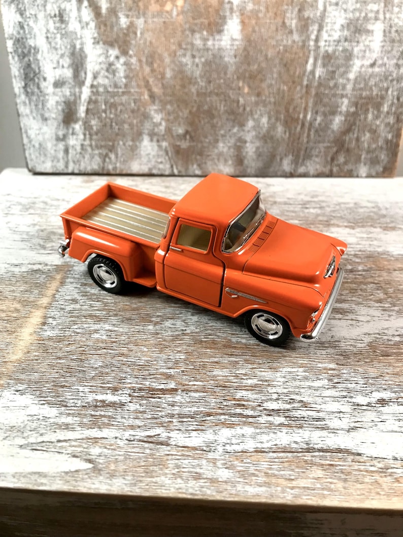 May include: An orange vintage pickup truck model with silver accents. The truck has a detailed design, including a bed with silver stripes and chrome wheels. The model is a classic collectible.
