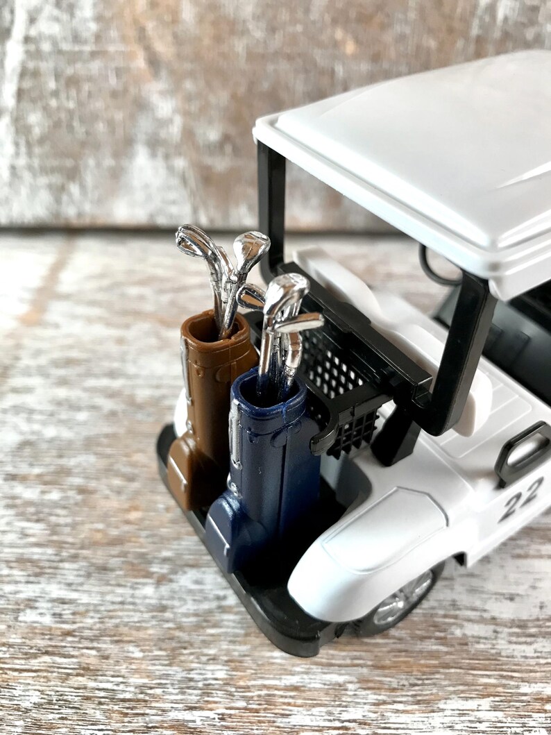 May include: A miniature white golf cart with a black frame and a number 22 on the side. The cart features a brown and blue golf bag with silver golf clubs. The cart is on a wooden surface.