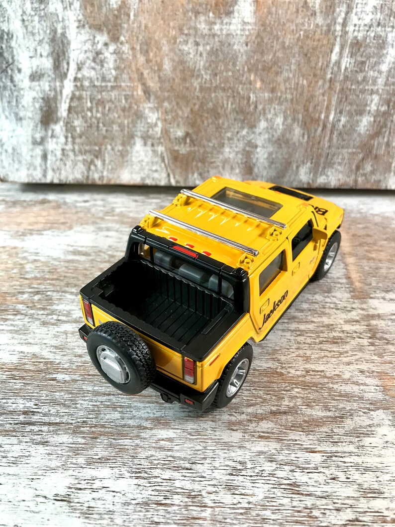 May include: Yellow toy truck with a black bed and a silver roof rack. The truck has a black spare tire on the back and the word "Jackson" written on the side.