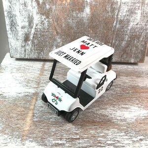 May include: A white toy golf cart with black accents, featuring the text "JUST MARRIED" and names "Matt" and "Jenn" with a red heart. The cart has the number "22" on the side and the word "4EVER" on the front.