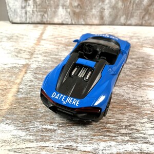 May include: A blue toy convertible car with a black engine cover. The car has the text "DATE HERE" in white on the back. The car is on a wooden surface.