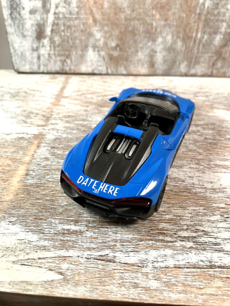 May include: A blue toy convertible car with a black engine cover. The car has the text "DATE HERE" in white on the back. The car is on a wooden surface.