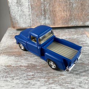 May include: A die-cast model of a classic blue pickup truck. The truck features a detailed interior, chrome accents, and a tan bed. The vehicle is a vintage style, with a glossy finish and realistic design.