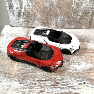 May include: Two toy sports cars, one red and one white, are displayed on a distressed wooden surface. The red car has black accents and a black interior. The white car has black accents and a black interior. Both cars have a sleek, modern design.