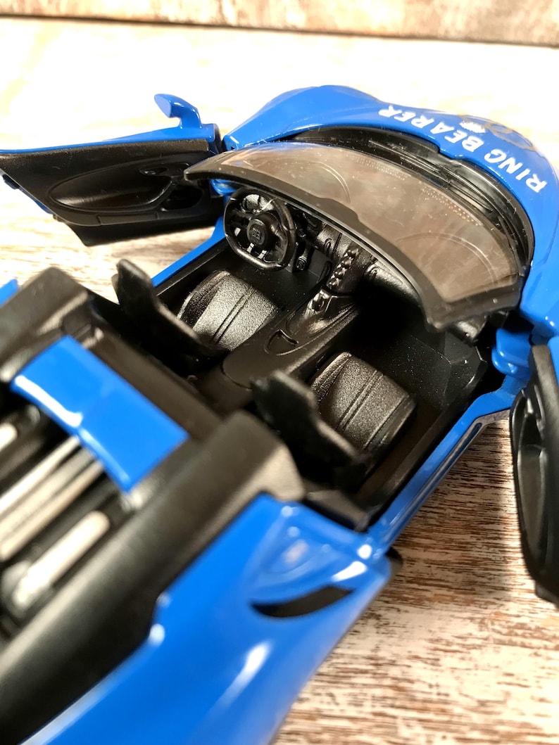 May include: A blue toy car with the doors open, revealing a black interior. The car has the words "RING BEARER" printed on the top. The steering wheel and dashboard are visible.
