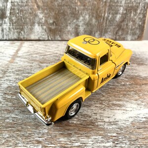 May include: A yellow toy truck with the text "Ring Bearer" and "Archie" printed on it. The truck has black tires and a silver bumper. The date "03-29-25" is visible on the windshield, along with two wedding rings.