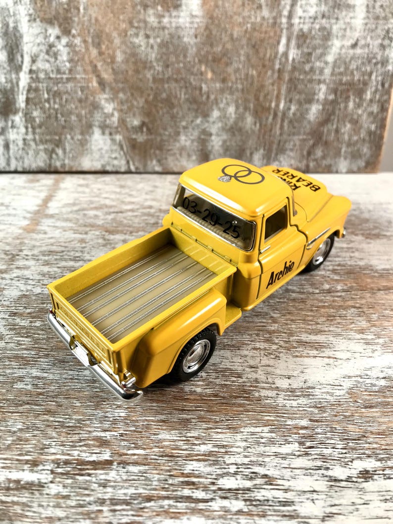 May include: A yellow toy truck with the text "Ring Bearer" and "Archie" printed on it. The truck has black tires and a silver bumper. The date "03-29-25" is visible on the windshield, along with two wedding rings.