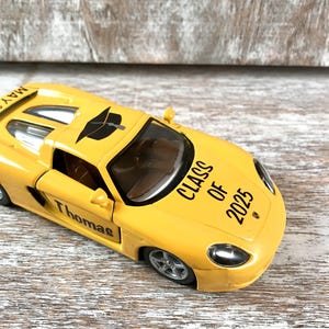 Puede incluir: Un coche de juguete amarillo con el nombre "Thomas" en la puerta y "Class of 2025" en el cap&oacute;. El coche tiene un birrete negro en el techo y la palabra "May" en el aler&oacute;n.