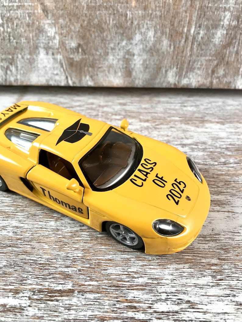 Puede incluir: Un coche de juguete amarillo con el nombre "Thomas" en la puerta y "Class of 2025" en el cap&oacute;. El coche tiene un birrete negro en el techo y la palabra "May" en el aler&oacute;n.