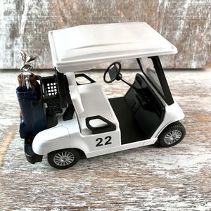 May include: A white toy golf cart with a black interior, a black steering wheel, and the number 22 on the side. The cart has a blue golf bag with golf clubs in the back. The cart has a white roof and black wheels.
