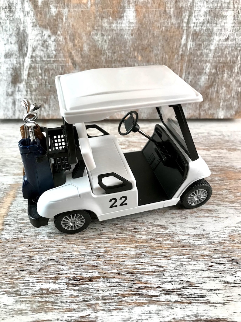 May include: A white toy golf cart with a black interior, a black steering wheel, and the number 22 on the side. The cart has a blue golf bag with golf clubs in the back. The cart has a white roof and black wheels.