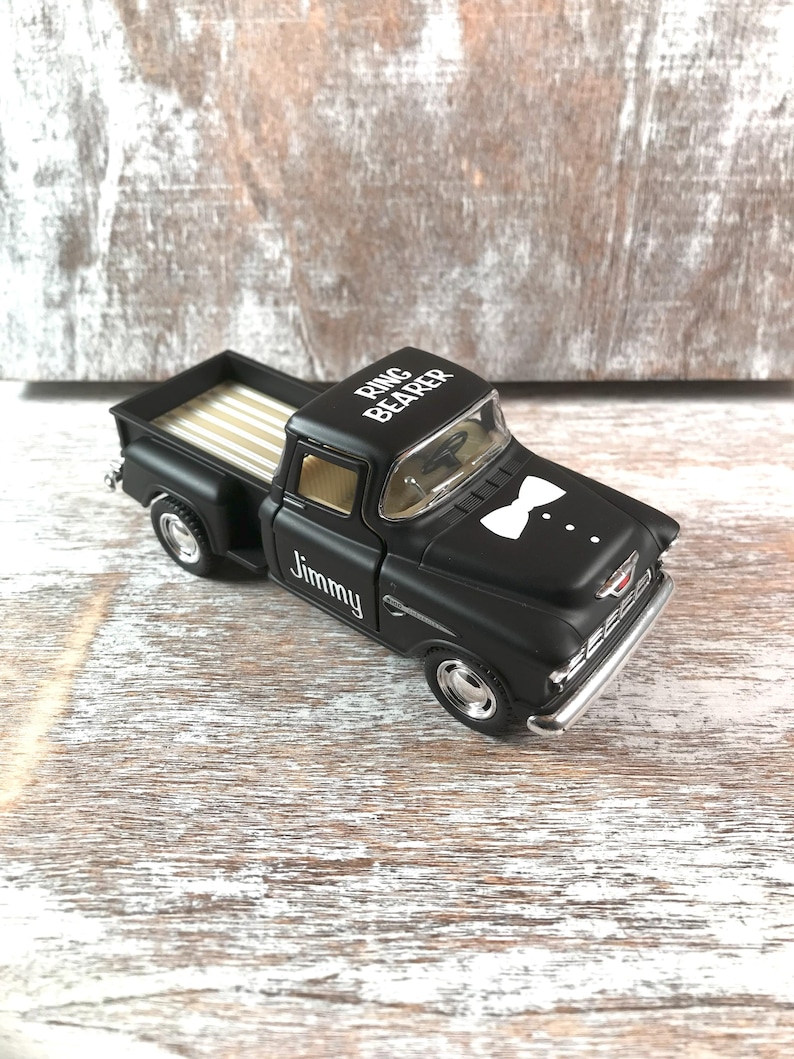 May include: A black toy pickup truck with the words "Ring Bearer" and "Jimmy" printed on it. The truck has a white bow tie graphic on the front and a tan bed. The truck is on a wooden surface.