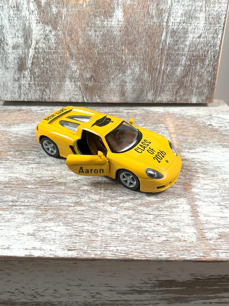 May include: A yellow toy car with the door open, displaying the name "Aaron". The car has the text "Class of 2026" on the hood and "Date Here" on the roof. The car is sitting on a light-colored wooden surface.