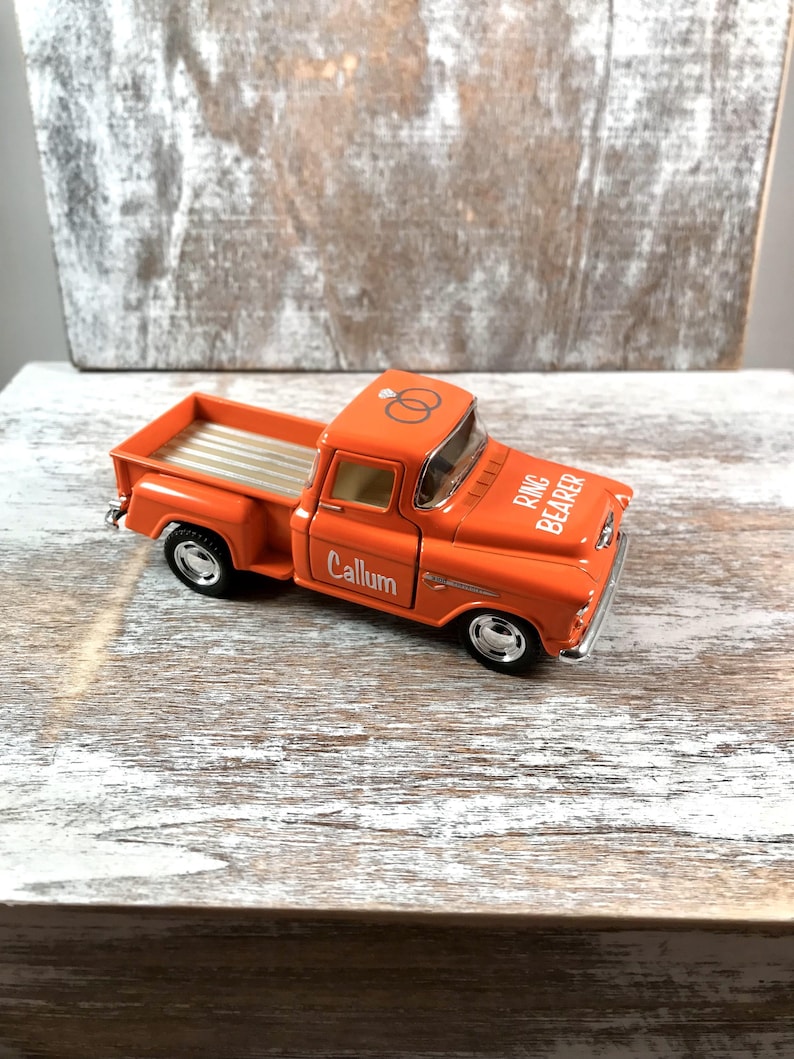 May include: Orange toy pickup truck with the words "Ring Bearer" and "Callum" printed on it. The truck has a silver bed and two wedding rings on the roof. The truck is a vintage style.