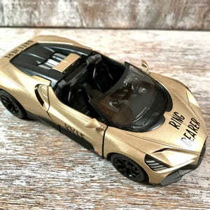 May include: A gold toy convertible car with black wheels and interior. The car has the words "RING BEARER" printed on the front and "DATE HERE" on the back. The car is sitting on a wooden surface.