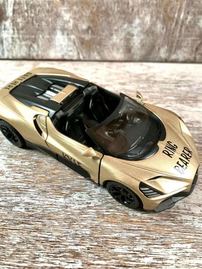 May include: A gold toy convertible car with black wheels and interior. The car has the words "RING BEARER" printed on the front and "DATE HERE" on the back. The car is sitting on a wooden surface.