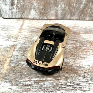 May include: A gold-colored toy sports car with a black engine cover and interior. The text "DATE HERE" is printed in red on the rear of the car. The car is sitting on a light-colored wooden surface.