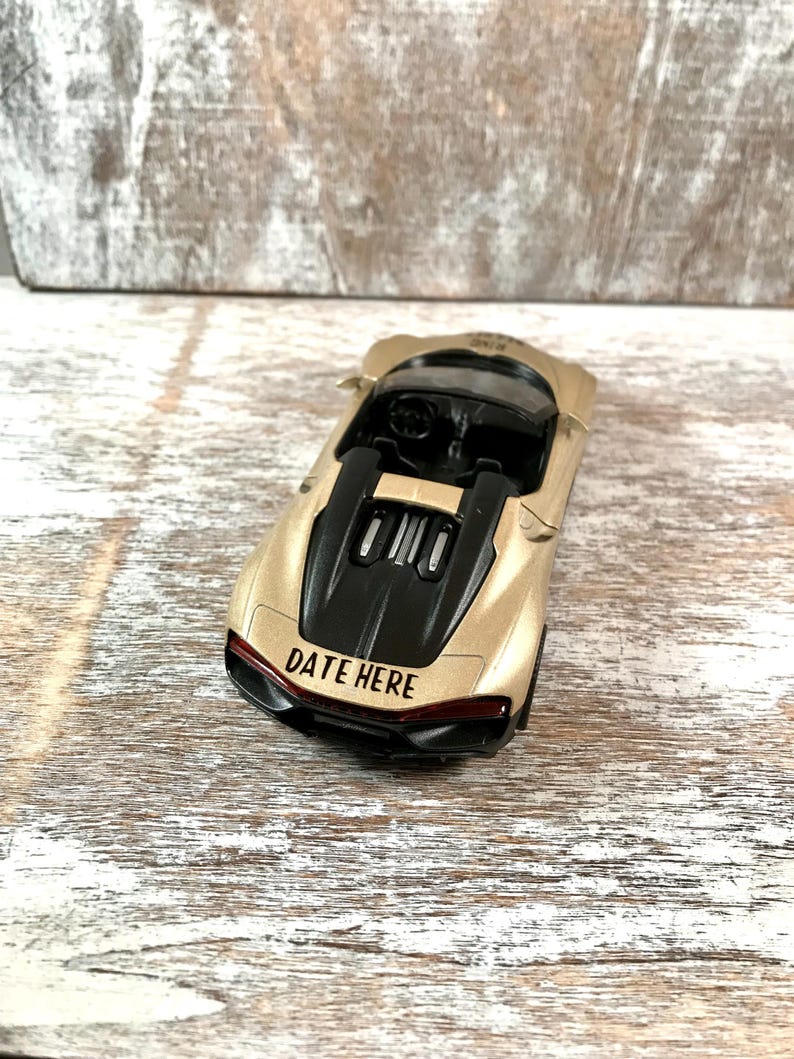 May include: A gold-colored toy sports car with a black engine cover and interior. The text "DATE HERE" is printed in red on the rear of the car. The car is sitting on a light-colored wooden surface.