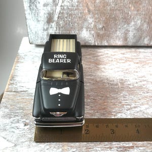May include: A black toy truck with the words "RING BEARER" on the hood and a white bow tie design. The truck bed has a light-colored wood-like pattern. A ruler shows the truck is approximately 4 inches long.