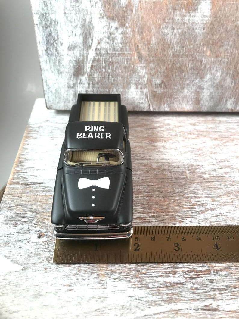 May include: A black toy truck with the words "RING BEARER" on the hood and a white bow tie design. The truck bed has a light-colored wood-like pattern. A ruler shows the truck is approximately 4 inches long.