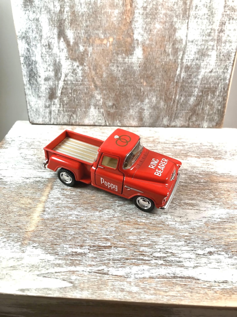 May include: A red toy pickup truck with silver wheels and a tan bed. The truck has the word "Poppy" on the door and "Ring Bearer" on the hood. Two gold wedding rings are on the roof.