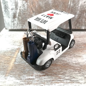 May include: A white golf cart model with black accents, carrying golf clubs and bags. The cart's roof displays "DATE HERE" and "JUST MARRIED" with names and a red heart. The number "22" is on the side.