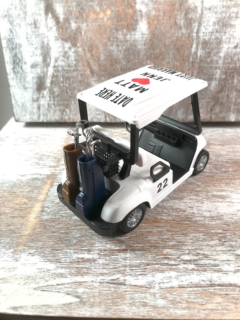 May include: A white golf cart model with black accents, carrying golf clubs and bags. The cart's roof displays "DATE HERE" and "JUST MARRIED" with names and a red heart. The number "22" is on the side.