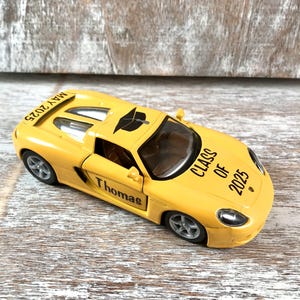 Puede incluir: Un coche de juguete amarillo con el nombre "Thomas" en el lateral y "Class of 2025" impreso en el cap&oacute;. El coche tiene un gorro de graduaci&oacute;n encima y la fecha "Mayo 2025" est&aacute; impresa en la parte trasera.