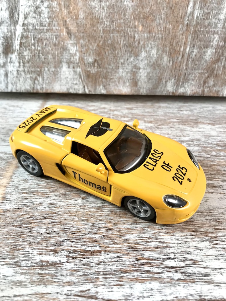 Puede incluir: Un coche de juguete amarillo con el nombre "Thomas" en el lateral y "Class of 2025" impreso en el cap&oacute;. El coche tiene un gorro de graduaci&oacute;n encima y la fecha "Mayo 2025" est&aacute; impresa en la parte trasera.