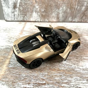 May include: A gold-colored toy car with black accents, including the wheels and interior. The doors are open, revealing the interior details. The text "DATE HERE" is visible on the back of the car. The car is sitting on a wooden surface.
