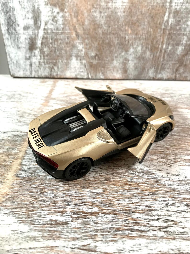 May include: A gold-colored toy car with black accents, including the wheels and interior. The doors are open, revealing the interior details. The text "DATE HERE" is visible on the back of the car. The car is sitting on a wooden surface.