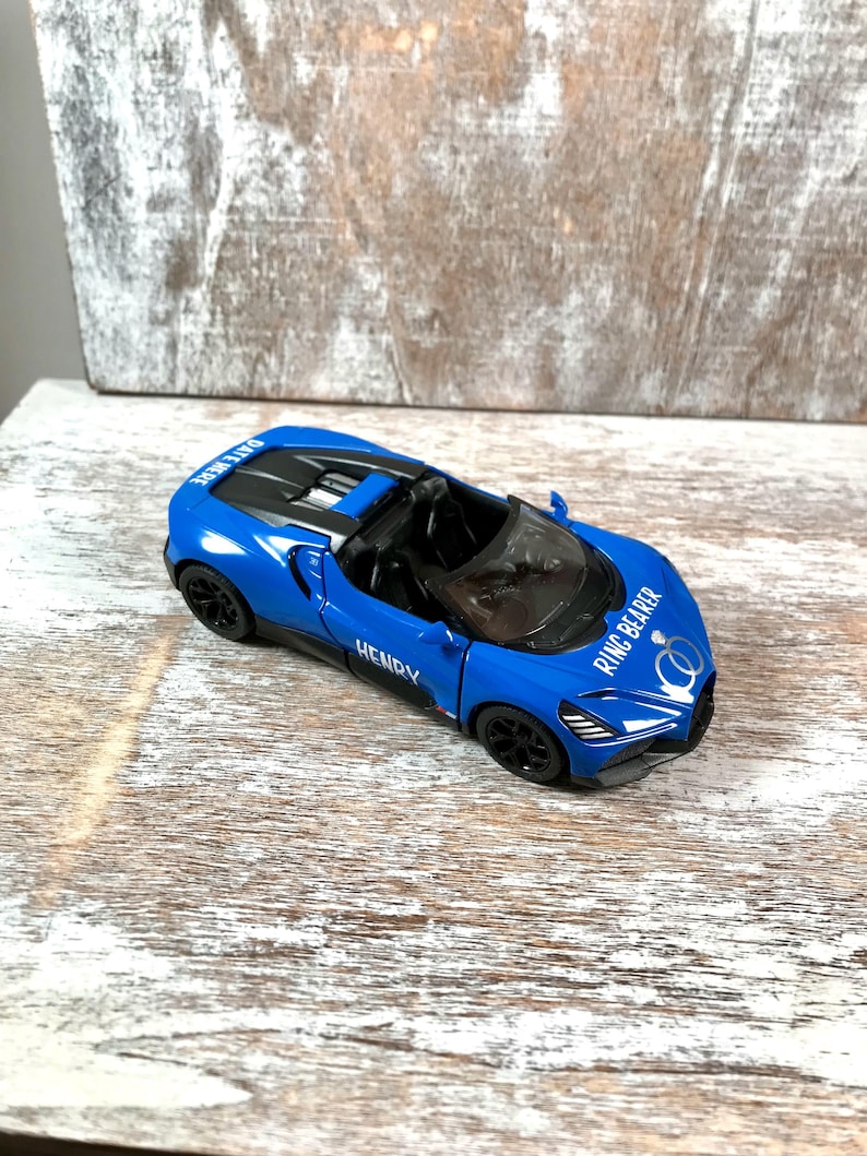 May include: A blue toy convertible car with black wheels. The car has the text "HENRY" on the side and "RING BEARER" on the hood. The car also has the text "DATE HERE" on the top.