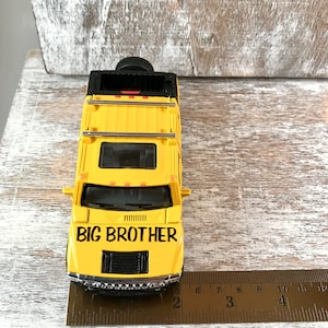May include: Yellow toy Hummer with black details and the words "BIG BROTHER" on the hood. The toy car is sitting on a wooden surface with a ruler next to it.